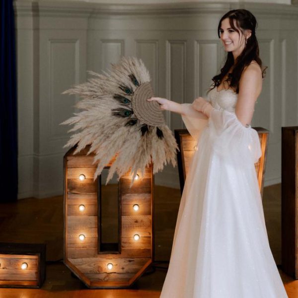 Braut im Brautkleid vor XXL-Love Lettern mit Pampasgrasfächer in der Hand.