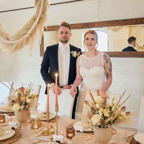 Brautpaar lacht in die Kamera vor einem dekorierten Hochzeitstisch in Naturtönen.